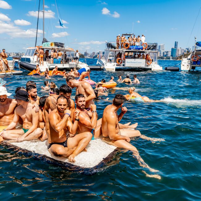 A group of men in swim trunks sits closely together on a floating platform in a busy bay filled with boats and people swimming. The sun is shining, and the atmosphere is lively, with the city skyline visible in the background. The Yacht Social Club Event Boat Charters hosts this social gathering on the water.