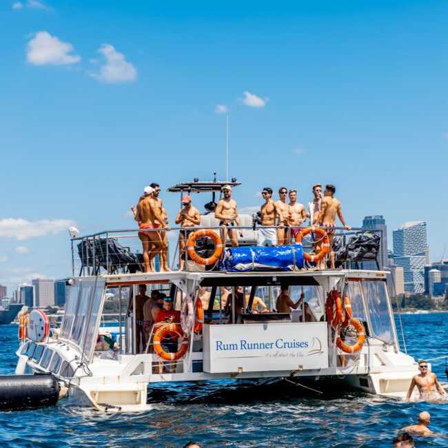 A group of people on a two-story boat labeled "Rum Runner Cruises" enjoys a sunny day on the water. The boat, a part of The Yacht Social Club Sydney Boat Hire, is equipped with lifebuoys and some passengers dip in the water or sunbathe on the deck. The skyline of a city with modern buildings is visible in the background.