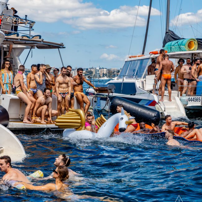 A lively scene at The Yacht Social Club Sydney Boat Hire shows numerous people in swimsuits socializing on deck and in the water. Some are on inflatables, and others are diving into the blue sea. In the background, other boats and a city skyline are visible under a clear, sunny sky.