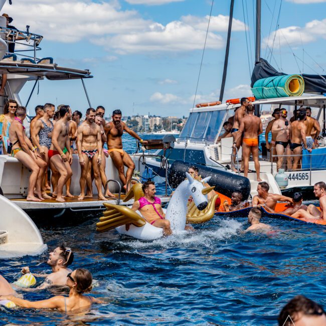 A lively Yacht Social Club event features people in swimwear on and around yachts. Some are dancing while others float in the water. A man rides an inflatable unicorn. The sky, filled with fluffy clouds, serves as a backdrop to the scene, creating a vibrant atmosphere.