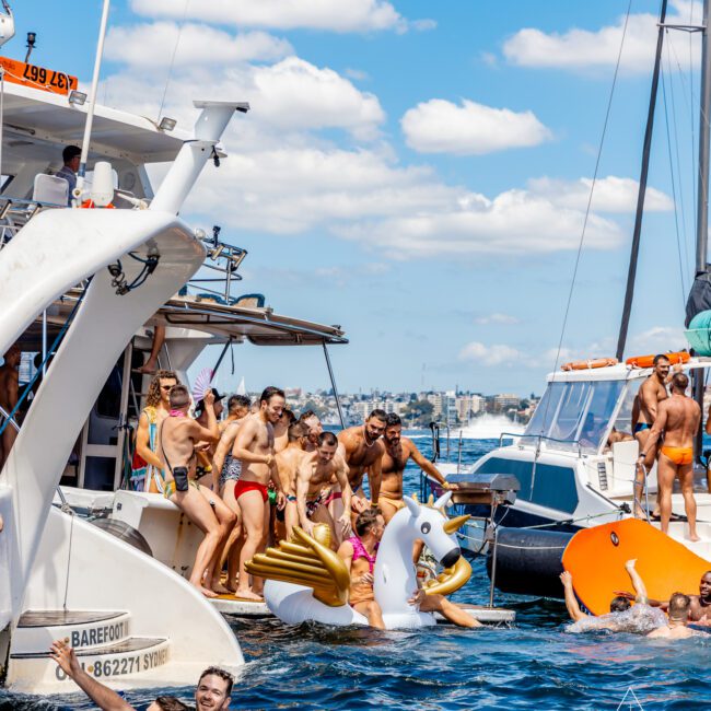 A lively boat party scene on a sunny day with people on and around several yachts, some playing and swimming in the water. A large inflatable unicorn float is in the water, and many attendees are enjoying The Yacht Social Club Sydney Boat Hire festivities.