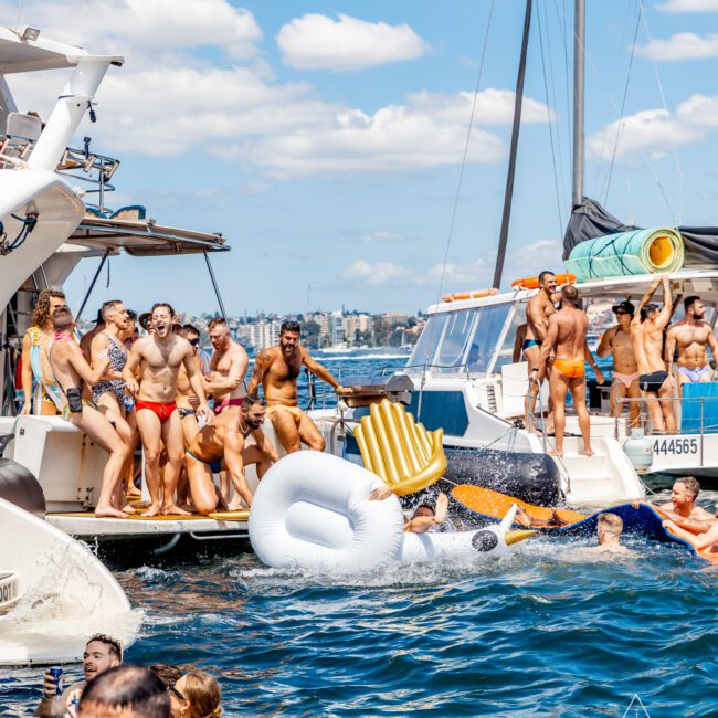 A lively gathering of people in swimsuits on docked boats and inflatables, enjoying a sunny day on the water at The Yacht Social Club Event. Some are diving in, while others socialize on the boats under a partly cloudy sky. In the background, city buildings are visible.