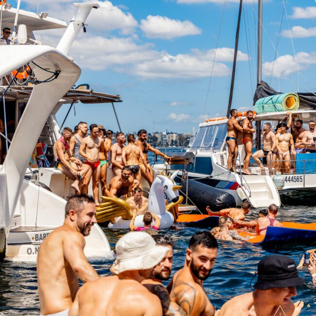 A large group of people are enjoying a sunny day on The Yacht Social Club Event Boat Charters in Sydney. Some are swimming, while others lounge on inflatable rafts and boat decks. Many are wearing swimwear, and the atmosphere is festive and lively under a bright blue sky.