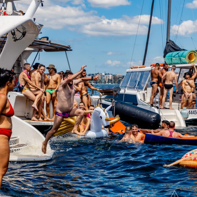 People are enjoying a day on the water, with some swimming and others on yachts from The Yacht Social Club Sydney Boat Hire. A man in colorful swim trunks jumps off a boat, while others watch or relax on inflatables shaped like a unicorn and other fun designs. The sky is clear with scattered clouds.