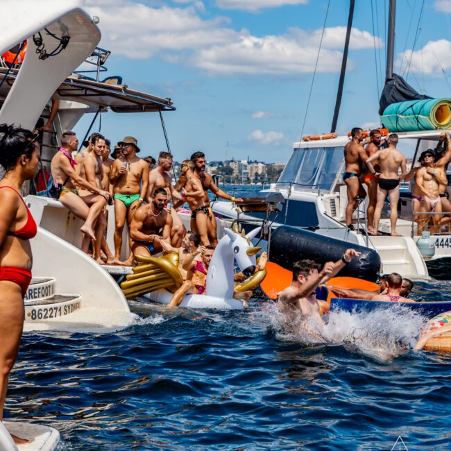 A lively scene of people on and around boats on a sunny day. Some are on the boat's deck, wearing swimwear and chatting, while others are in the water swimming or relaxing on inflatables, including a shark float. The sky is blue with a few clouds. Experience it all with Sydney Harbour Boat Hire The Yacht Social Club.