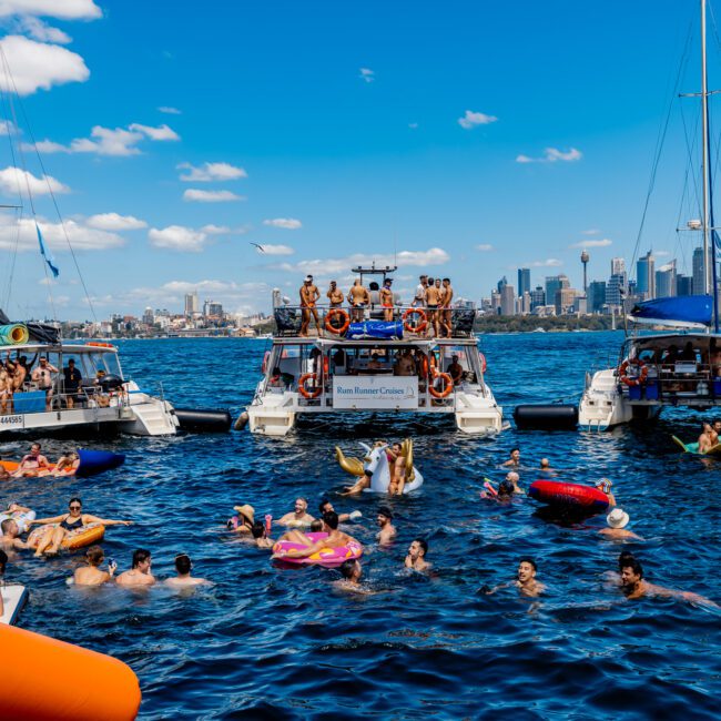 A lively scene on the water with people enjoying a sunny day. Several boats from The Yacht Social Club Event Boat Charters are docked together, and many individuals are swimming, floating on inflatables, and socializing. The city skyline is visible in the background under a blue, partly cloudy sky. Logos include GAYM and Yacht Social.