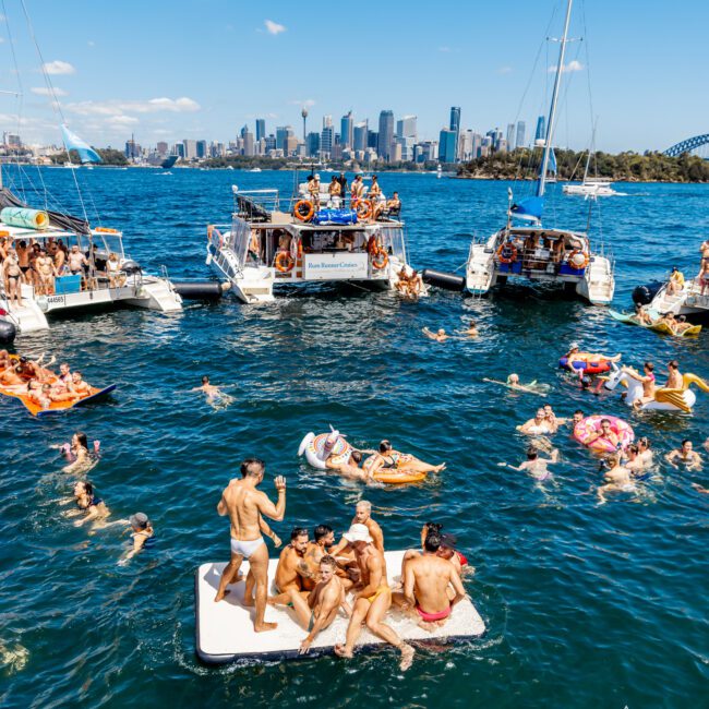 A lively scene of people enjoying a sunny day on the water. Various individuals are seen swimming and floating on inflatable devices between several boats. In the background is a city skyline with tall buildings and a clear blue sky, characteristic of Sydney Harbour Boat Hire The Yacht Social Club.