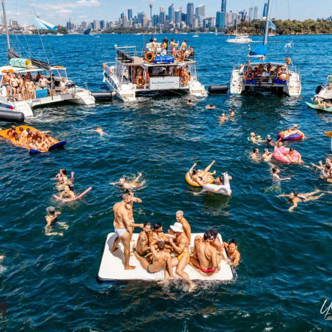 A group of people enjoying a sunny day on the water are seen on yachts and inflatables in a blue harbor. Some swim, while others relax on floating platforms. In the background, a city skyline and a bridge are visible. The Yacht Social Club Sydney Boat Hire adds to the lively, festive atmosphere.