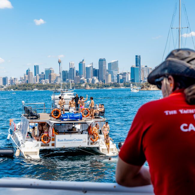 A man wearing a red shirt with "Captain" on the back looks out from a boat at other boats and people floating on blue water. The backdrop features the skyline of a modern city with tall buildings and the iconic Sydney Harbour Bridge visible on the right, part of The Yacht Social Club Event Boat Charters.