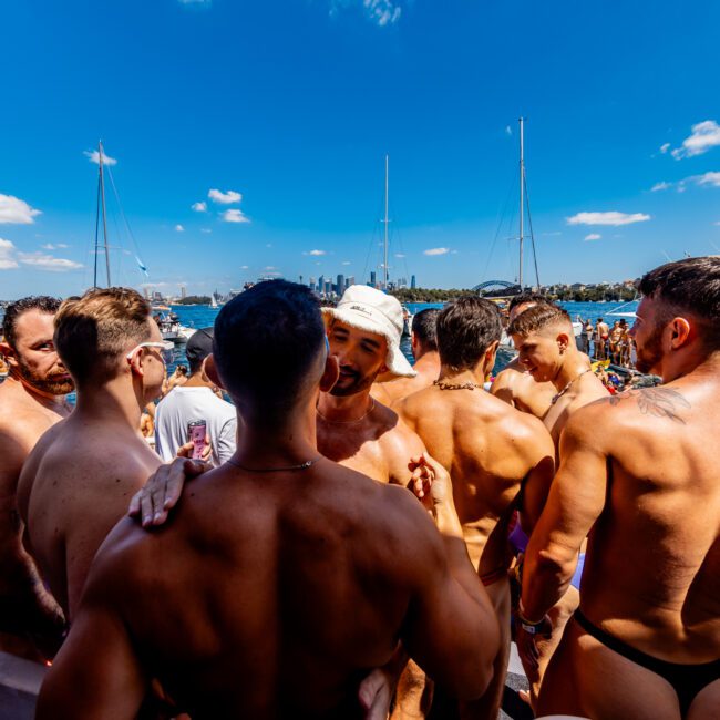 A group of shirtless men socialize on a boat under a clear blue sky, enjoying the luxury offerings of The Yacht Social Club Sydney Boat Hire. Other boats and people can be seen in the background on the water, enhancing the vibrant atmosphere of their sunny day out.