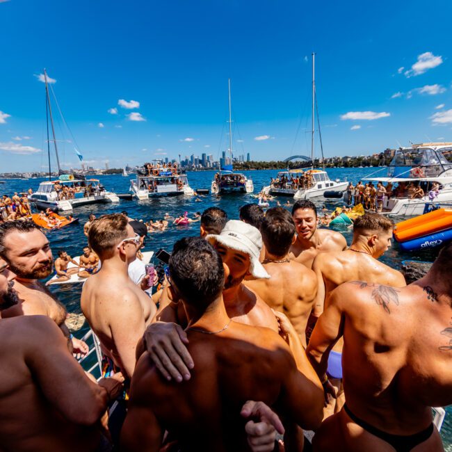 A large group of shirtless individuals are gathered on a boat under a bright blue sky, with more people on other boats nearby. The scene depicts a lively party atmosphere on the water. Sydney’s skyline, including the Harbour Bridge, is visible in the background during The Yacht Social Club Event Boat Charters.