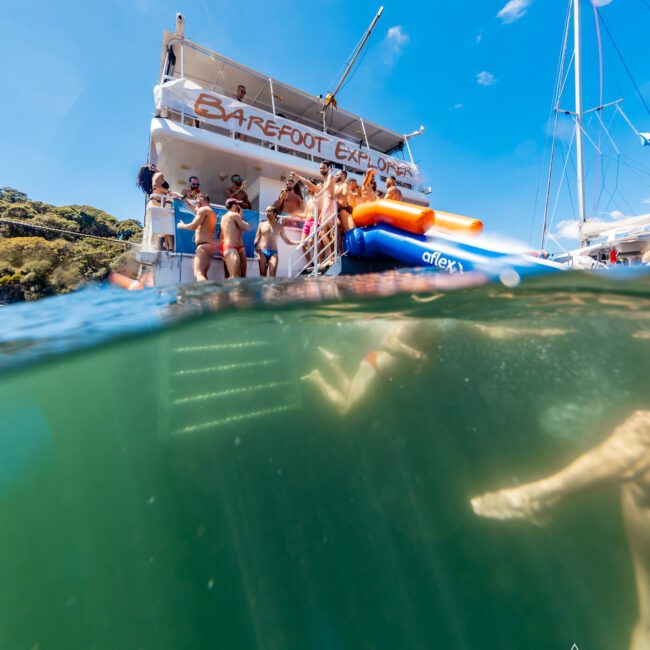 People are gathered on the deck of a boat named "BAREFOOT EXPLORER," with several standing near an inflatable slide that descends into the water. The lower half shows an underwater view of legs and feet near the surface, capturing the lively vibe of a Sydney Harbour Boat Hire event by The Yacht Social Club.