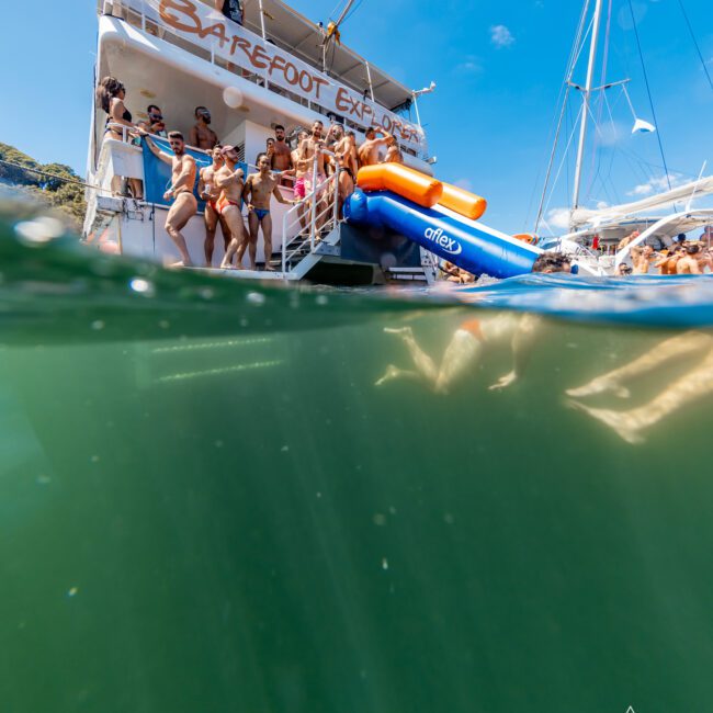 A group of people in swimwear stand on the deck of a boat named "Barefoot Explorer," part of The Yacht Social Club Sydney Boat Hire, preparing to slide down a large blue and orange waterslide into the sea. The bottom half of the image shows a view underwater, revealing greenish water. Clear skies are overhead.