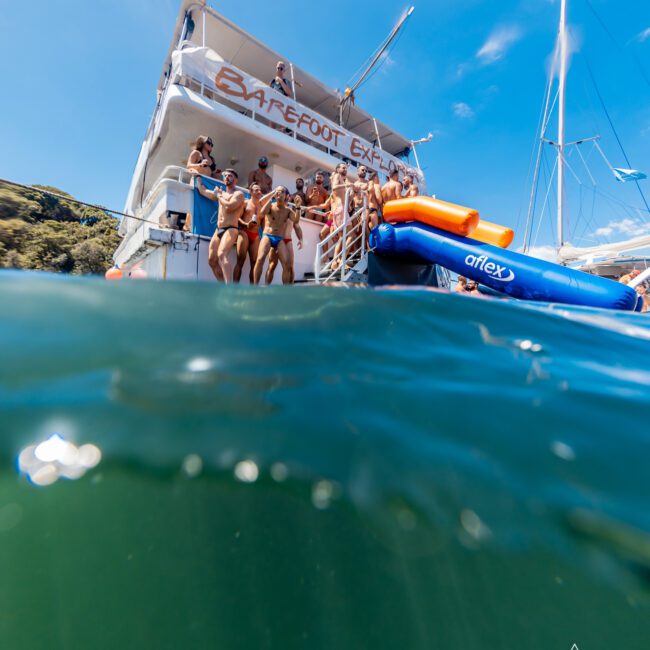 A group of people are diving and sliding off a boat with "Barefoot Explorer" signage into the sea. The photo captures a blend of underwater and above-water perspectives. Logos for "GAYM" and "The Yacht Social Club" are visible in the corners, highlighting Luxury Yacht Rentals Sydney.