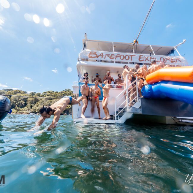 A group of people are enjoying a sunny day on a boat named "Barefoot Explorer" from The Yacht Social Club. Some are standing on the deck, while others are using slides that lead into the water. One person is diving in. The background shows trees and a clear blue sky, perfect for an exclusive event with Sydney Harbour Boat Hire.