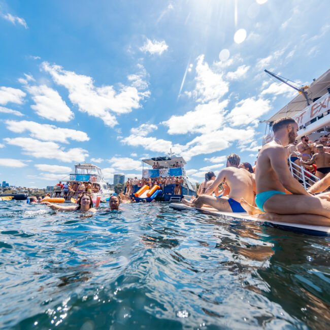 People enjoying a sunny day on the water and on boat decks. The scene includes individuals swimming, lounging, and socializing on various boats, including one with a slide. The sky is blue with scattered clouds. Logos of GAYM and The Yacht Social Club are visible at this lively Boat Parties Sydney event.