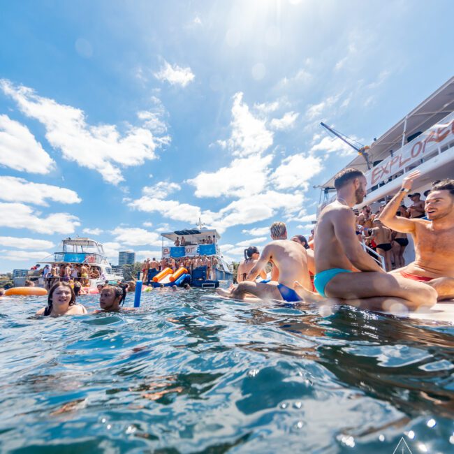 Group of people in swimsuits enjoying a sunny day on the water with multiple boats in the background. Some are swimming, while others sit and socialize on the boats. The blue sky is dotted with a few clouds. Logos for "GAYM" and "The Yacht Social Club" are visible. Boat Rental and Parties Sydney with The Yacht Social Club ensure a great time.