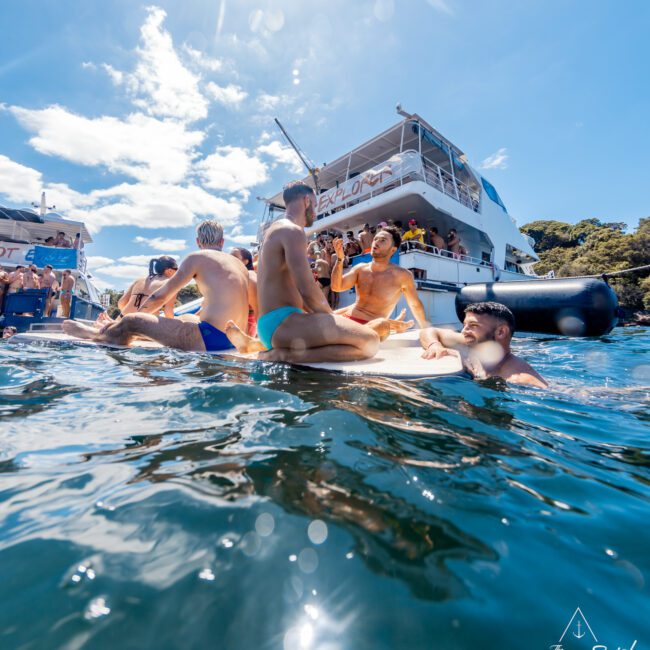 A group of people enjoy a sunny day on the water; some sit on an inflatable platform, others swim around. Behind them are two large yachts with more people on board from The Yacht Social Club Event Boat Charters. The sky is clear with a few clouds, making for a lively and social occasion.