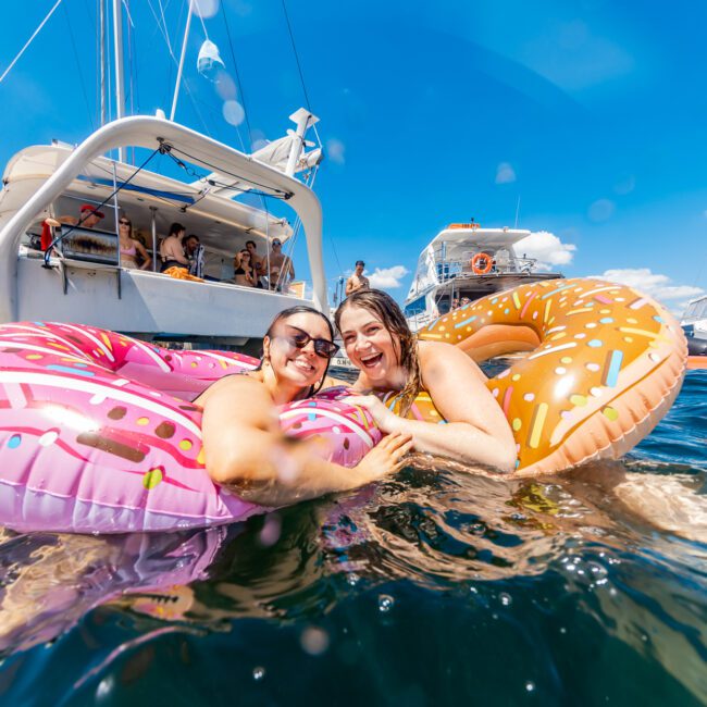 Two women float in a body of water on inflatable donut pool floats—one pink with sprinkles and one brown. In the background, several boats from The Yacht Social Club are anchored, filled with people. The sky is clear and blue, perfect for a day of boat parties in Sydney.