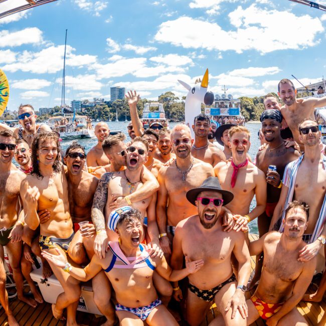 A joyful group of men and women in swimwear pose together on a luxury yacht rental during a sunny day. There are decorated boats and inflatables in the background, with more people enjoying the water. Everyone looks happy, smiling, and celebrating.