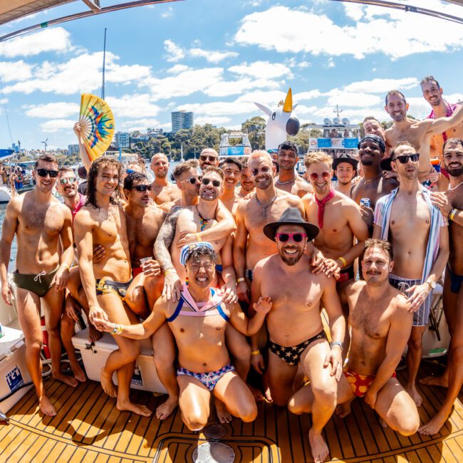 A large group of people in swimsuits is posing for a photo on the deck of a boat with a festive atmosphere. Other boats and partygoers are visible in the background, showcasing The Yacht Social Club Sydney Boat Hire. The weather is sunny, and everyone appears to be smiling and enjoying themselves.