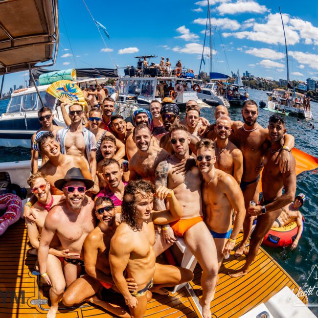 A large group of people in swimwear pose closely together aboard a boat on a sunny day. The background features another boat and a coastline under a clear blue sky. The mood is festive, and everyone is smiling and enjoying *The Yacht Social Club Event* hosted by *Luxury Yacht Rentals Sydney*.