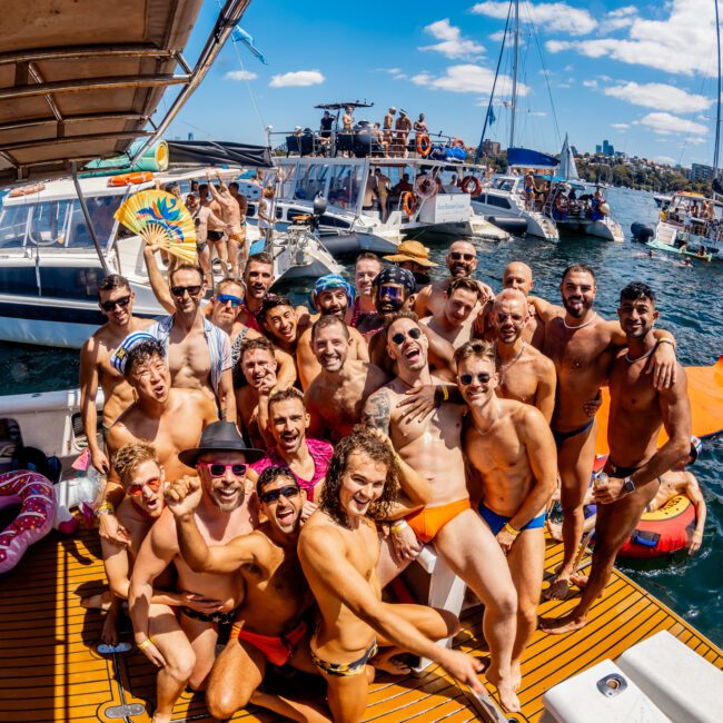 A joyful group of people, mostly in swimwear, pose together on the deck of a boat under a bright blue sky. Surrounding boats and the scenic Sydney Harbour enhance the lively atmosphere, making it clear this is a memorable Yacht Social Club event.