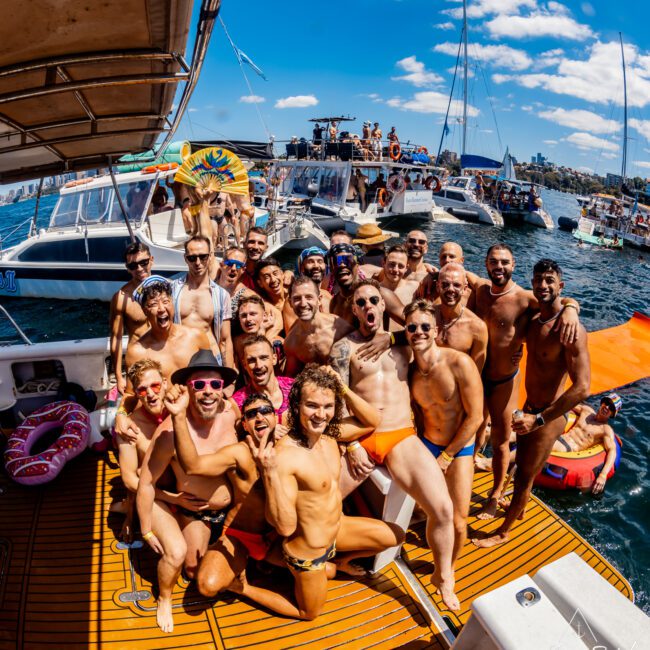 A lively group of friends, mostly men in swimwear, pose enthusiastically on a boat. The boat is surrounded by other boats from The Yacht Social Club on a sunny day against a clear blue sky. The atmosphere is festive, with people smiling, cheering, and enjoying the outing on the water.