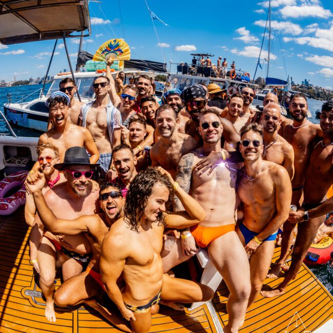 A large group of joyful people, mostly men in colorful swimwear, are posing together on a boat deck under a bright, sunny sky. They are smiling, laughing, and some are flexing their muscles, creating a lively, festive atmosphere during The Yacht Social Club Event Boat Charters. Another boat is visible in the background.