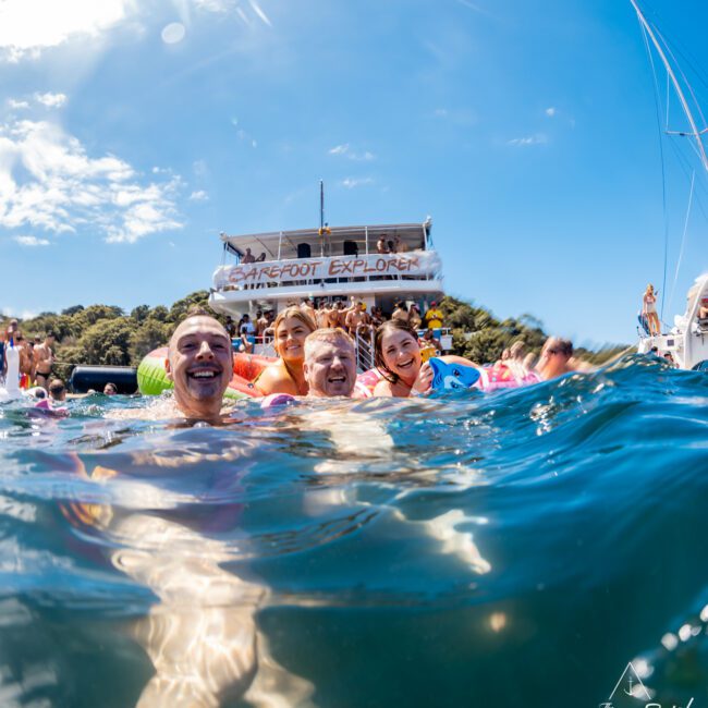 Group of people smiling and enjoying the water near a boat labeled "Barefoot Explorer" on a sunny day. Some are holding onto inflatable floats, with the boat and a scenic background of trees and blue sky visible, showcasing an ideal moment for The Yacht Social Club Sydney Boat Hire.