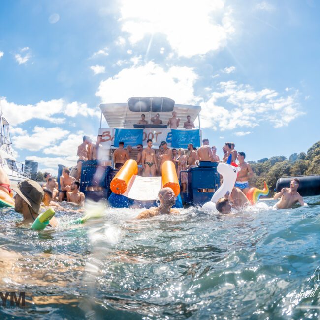 A lively scene of people enjoying a sunny day on a boat and in the water. Some are on a large Luxury Yacht Rentals Sydney with a slide, while others swim and play with inflatable toys in the water. The sky is clear, and there is lush greenery in the background.