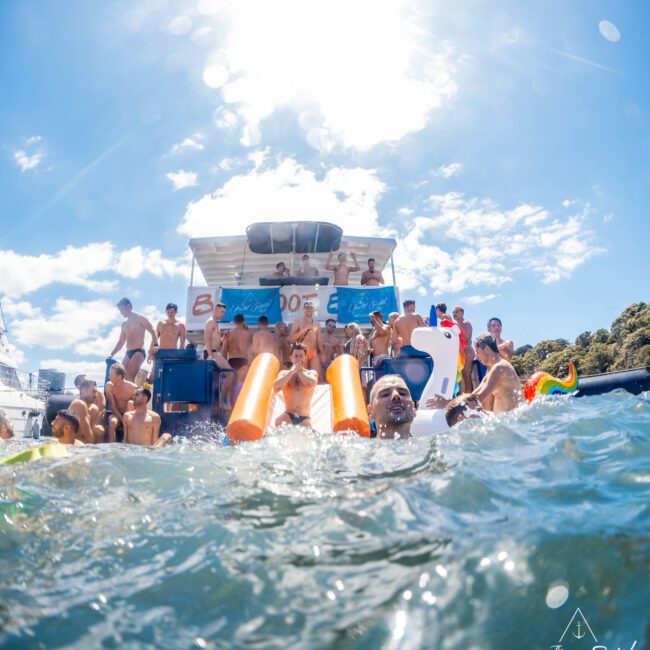 A vibrant scene of numerous individuals enjoying a sunny day on a yacht and in the water. People are socializing, some on inflatable floats, with clear blue skies above. Logos for "Yacht Social Club" and "GAYM" are visible. The atmosphere is lively and festive at Boat Parties Sydney The Yacht Social Club.