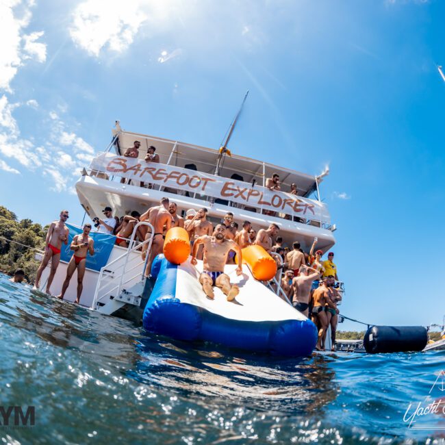 A large group of people enjoying a sunny day on a yacht named "Barefoot Explorer." Some use an inflatable slide to splash into the water, while others relax on the deck. The Yacht Social Club ensures the perfect setting for this Sydney Harbour boat hire, with clear blue skies and calm seas in the background.