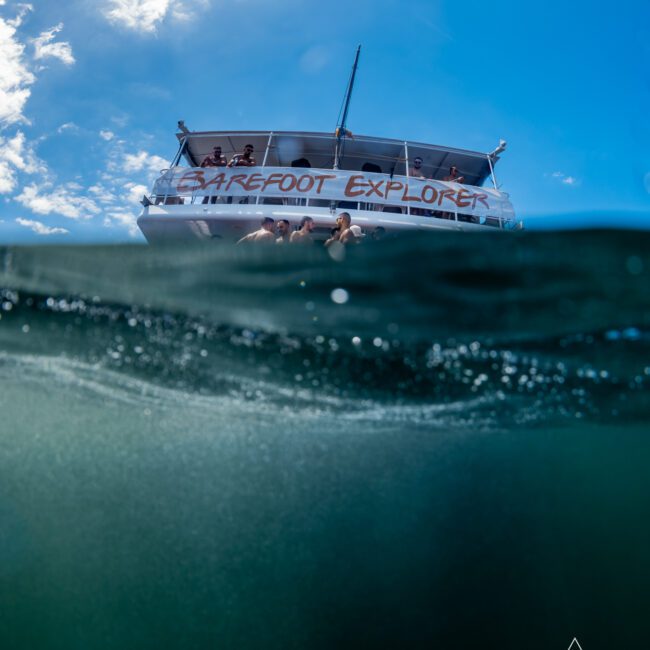 An image of a boat named "Barefoot Explorer" with people on board, partially submerged in water. The lower half shows an underwater view. Logos for GAYM and The Yacht Social Club Event Boat Charters are visible at the bottom corners. Sky is clear with some clouds.