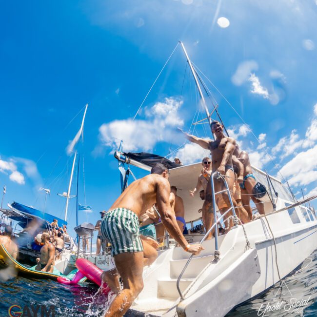 A group of people enjoying a sunny day on a boat. Some are on the deck, and one person is climbing up a ladder from the water. The sky is clear and blue, the scene is lively with other boats in the background, showcasing a perfect gathering by The Yacht Social Club Event Boat Charters.