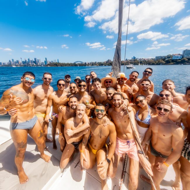 A large group of smiling people dressed in swimwear, gathered on a boat on a sunny day. They are posing closely together with the water and a skyline of buildings in the background. The atmosphere appears festive and joyful, capturing the essence of Sydney Harbour Boat Hire The Yacht Social Club.