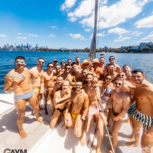 A group of men in swimwear are gathered on a sailboat on a sunny day with a cityscape in the background. They are smiling and posing for the camera. The image has logos for "GAYM" and "The Yacht Social Club Event Boat Charters" at the bottom corners, showcasing the fun of Boat Parties Sydney.