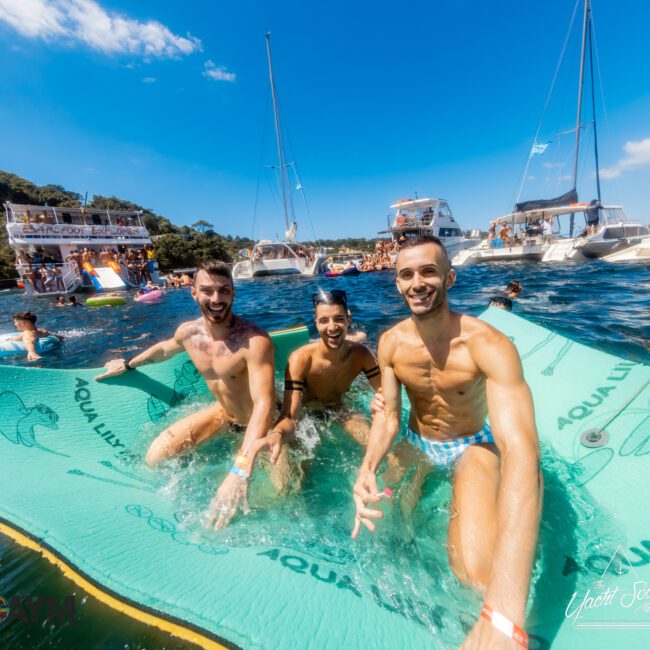 Three men are smiling and posing for a photo while sitting partially submerged on a floating mat in the ocean. In the background, several yachts from The Yacht Social Club are anchored nearby with people socializing on board and in the water. The sky is clear and sunny.