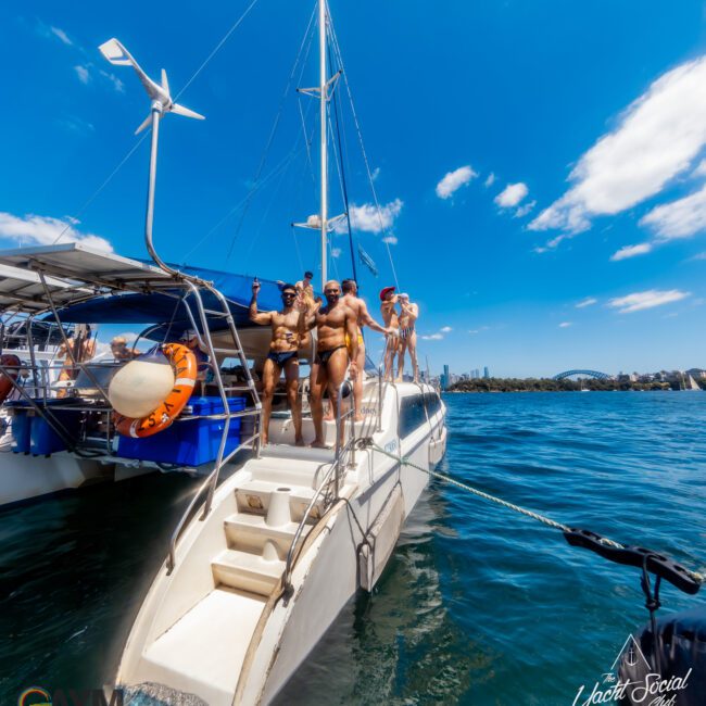 A group of people in swimwear are standing on the deck of "The Yacht Social Club" anchored in a body of water. They appear to be enjoying sunny weather with a cityscape and blue sky in the background. Experience premier Boat Parties Sydney through The Yacht Social Club Event Boat Charters.