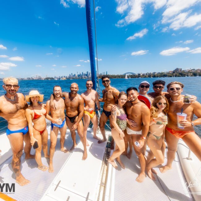 A group of people in colorful swimwear are smiling and posing on a boat with water and a city skyline in the background. The sky is sunny and blue, perfect for The Yacht Social Club Event Boat Charters. Logos for "GAYM" and "Hainsley" are visible at the bottom corners.