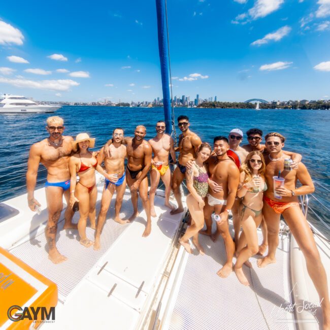 A group of people in swimsuits stand together on a boat under a sunny sky with a city skyline and bridge in the background. They appear to be having fun, smiling, and some are holding drinks. The image features logos for "GAYM" and "GAY Water Sports," promoting Sydney Harbour Boat Hire The Yacht Social Club.