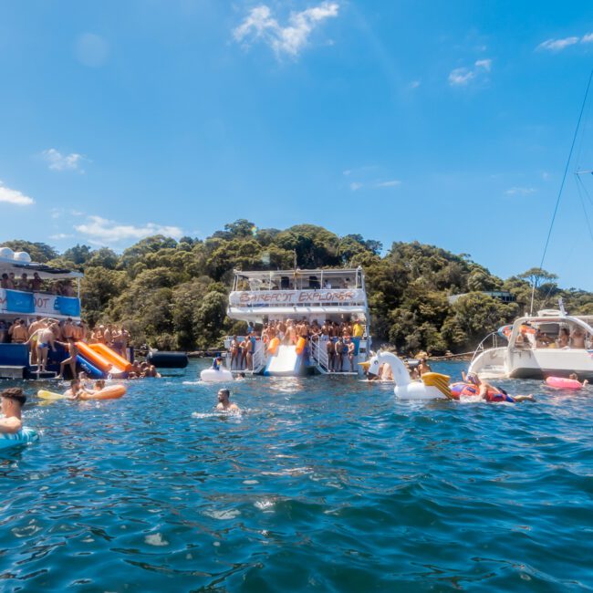 A lively boat party takes place on a sunny day, with several boats docked closely together on a beautiful blue sea. People are enjoying themselves on inflatables in the water and on the boats, with a dense, green tree line in the background. Experience this joy with Luxury Yacht Rentals Sydney and The Yacht Social Club.