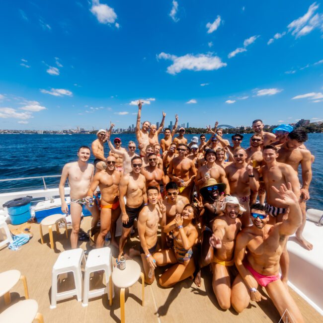 A large group of people on a boat from The Yacht Social Club, wearing swimwear and posing for a photo. Some are standing, others are sitting, with many raising their hands and smiling. The boat is on a body of water with a clear blue sky and distant shoreline visible in the background.