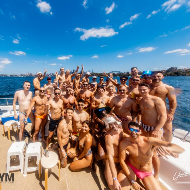 A large group of people in swimwear poses for a photo on a boat deck under a clear blue sky. Some are sitting while others stand, smiling and making peace signs. The setting suggests a festive, sunny day on the water, similar to events hosted by The Yacht Social Club Sydney Boat Hire.