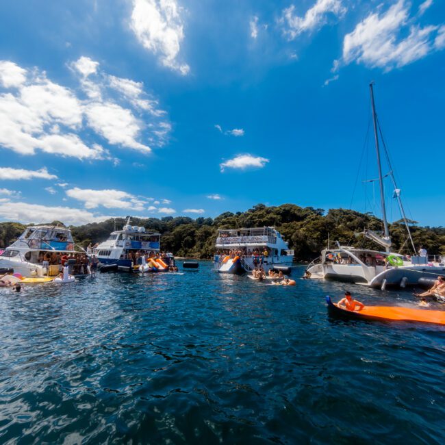 A vibrant outdoor scene with multiple boats and yachts on a sunny day, anchored near a lush green island. People are swimming, lounging on inflatables, and socializing on the boats. The sky is bright blue with scattered clouds. Experience the best with Luxury Yacht Rentals Sydney by The Yacht Social Club.