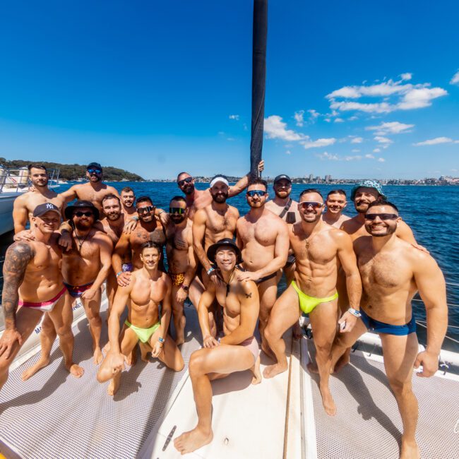 A group of smiling people in swimwear poses together on a boat deck under a sunny blue sky, enjoying The Yacht Social Club Event Boat Charters. They are on the water with a scenic view of the coastline and city skyline in the background. Some are standing while others are sitting or kneeling on the deck.