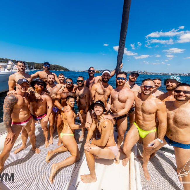 A group of shirtless men in swimsuits pose for a photo on a yacht under a clear blue sky, enjoying a sunny day with The Yacht Social Club. Sailboats and a coastline are visible in the background. Logos for AYM and Yacht Social Club are in the corners.