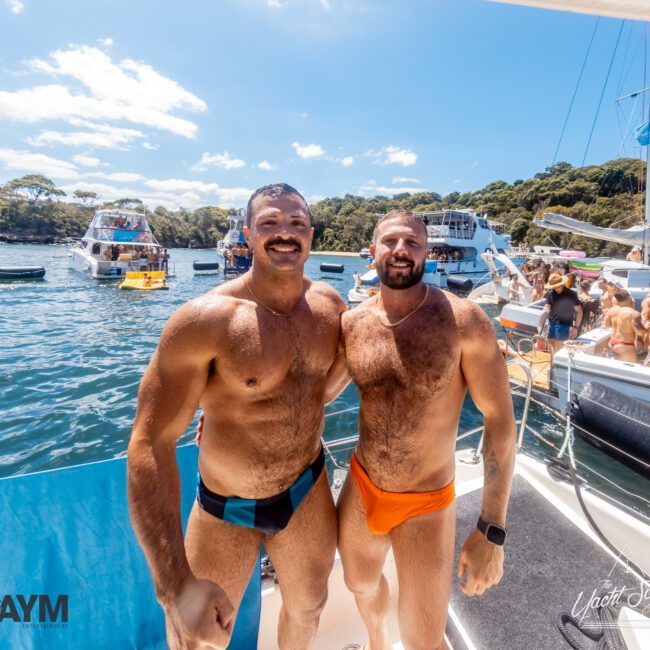 Two men in swimwear stand on a yacht deck, smiling at the camera. Other boats and people are visible in the background, enjoying a sunny day on the water with The Yacht Social Club. The scene suggests a lively and fun outdoor event. Logos for "GAYM" and "Yacht Social" are present in the corners.