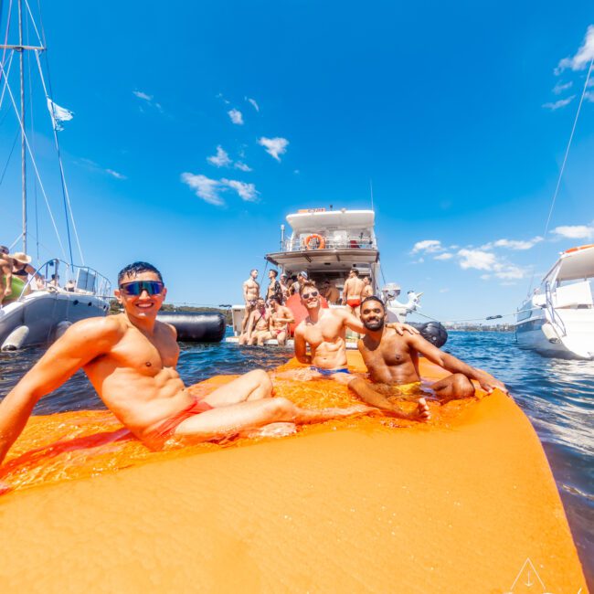 A group of men are lounging on an inflated orange float in the water, wearing swimsuits. Boats and more people are visible in the background on a sunny day with clear blue skies. Logos for "GAYM" and "The Yacht Social Club Sydney Boat Hire" are at the bottom corners of the image.