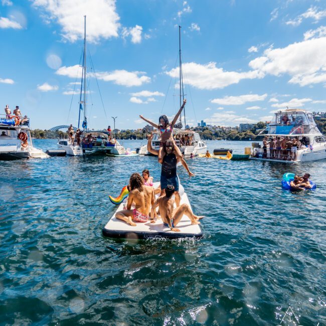 A group of people enjoying a sunny day on a small inflatable mat in the water surrounded by several boats. Some are sitting, while one person stands with arms raised. Others are swimming nearby. In the background, more people from The Yacht Social Club Sydney Boat Hire are on boats under a clear blue sky.